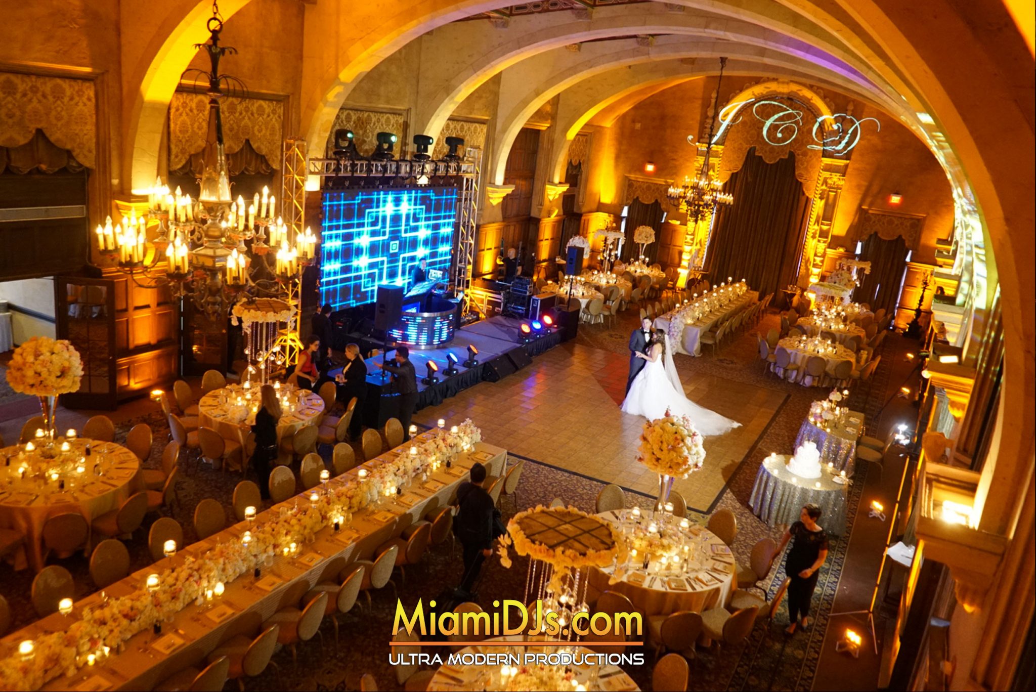A couple dancing alone in a gold-uplit ballroom with chandeliers and an LED wall behind them