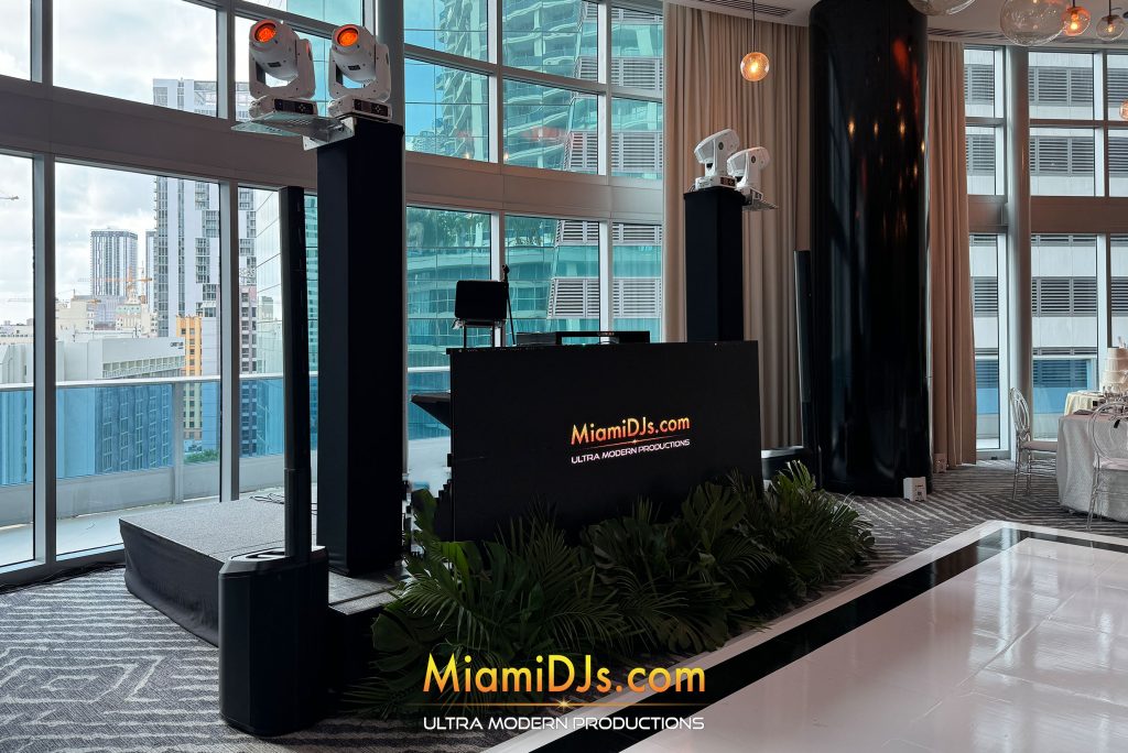 All-black column array speakers flanking a DJ booth against floor-to-ceiling windows